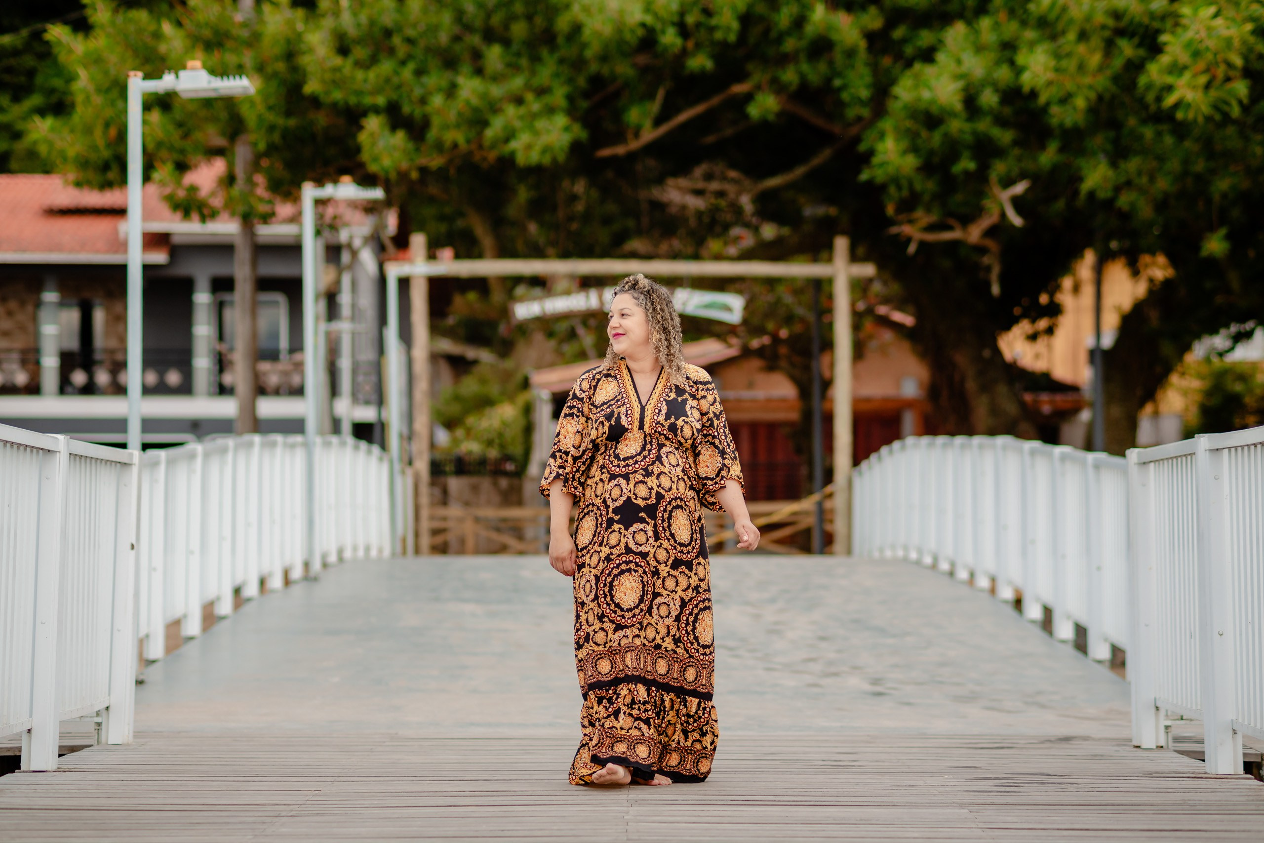 Fotógrafo ensaio gestante em Penha; retrato de corpo inteiro da gestante caminhando elegante sobre uma ponte de madeira em meio à natureza.