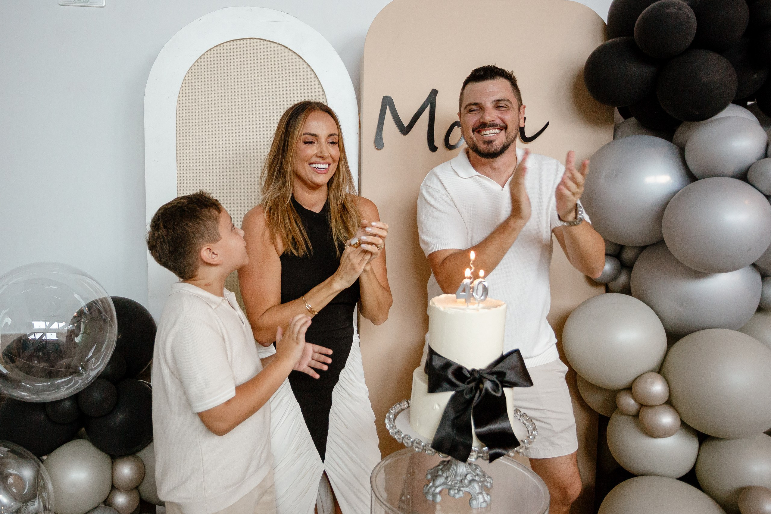 Fotografia de cobertura de festa de aniversário em Itajaí retratando a família sorridente diante do bolo festivo com velas acesas, enquanto o marido aplaude com entusiasmo.