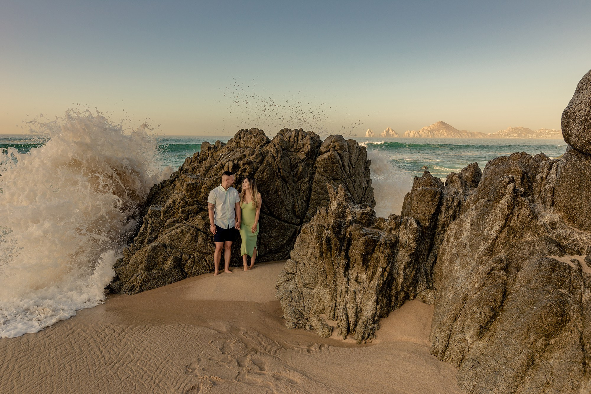 Couple portrait at Cabo San Lucas Arch – holding hands as ocean wave splashes against rocks, cinematic beach photography