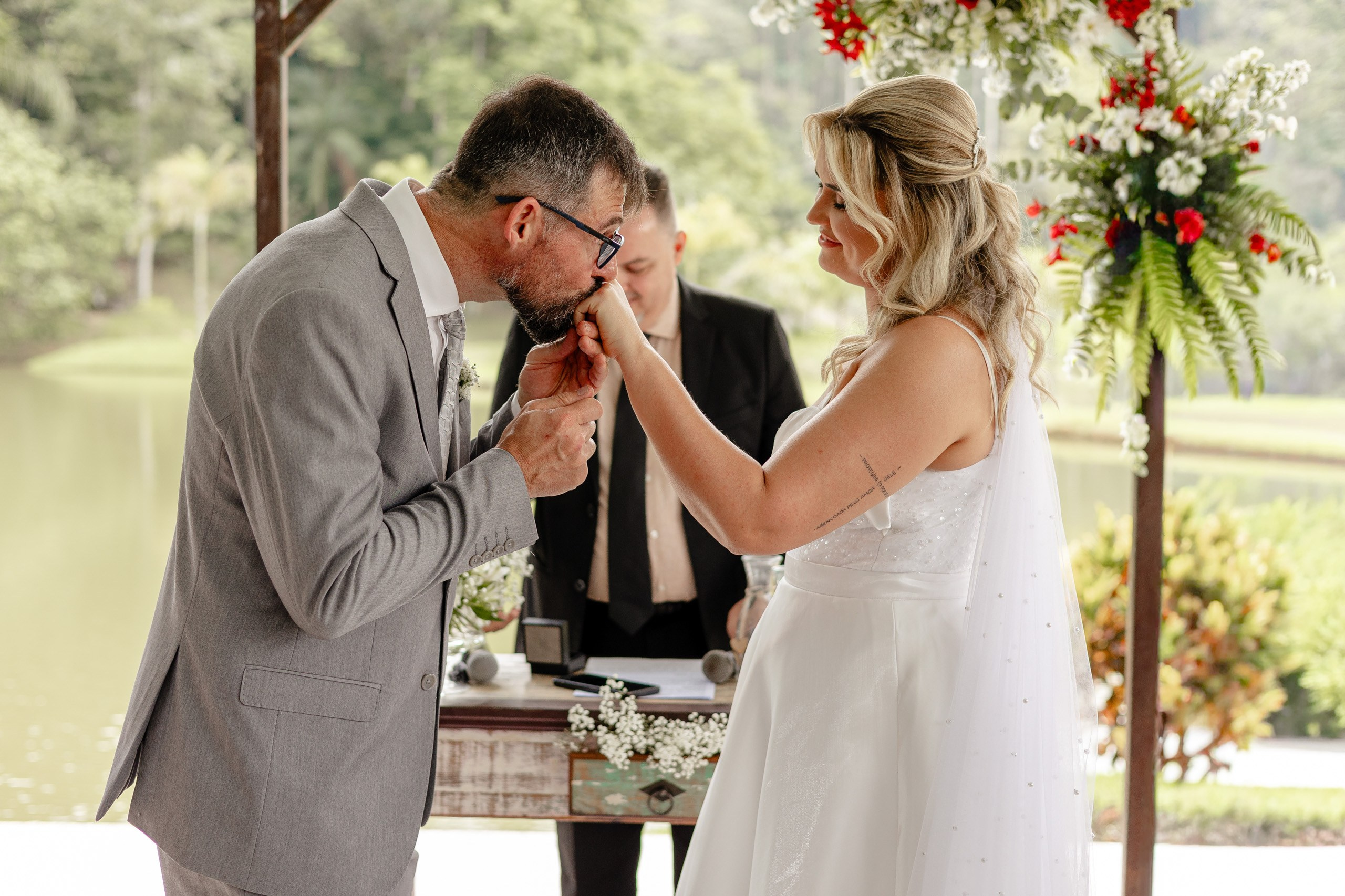 Fotógrafo de casamento em Santa Catarina; noivo beijando a mão da noiva no altar com decoração floral e paisagem natural ao fundo durante cerimônia no campo.