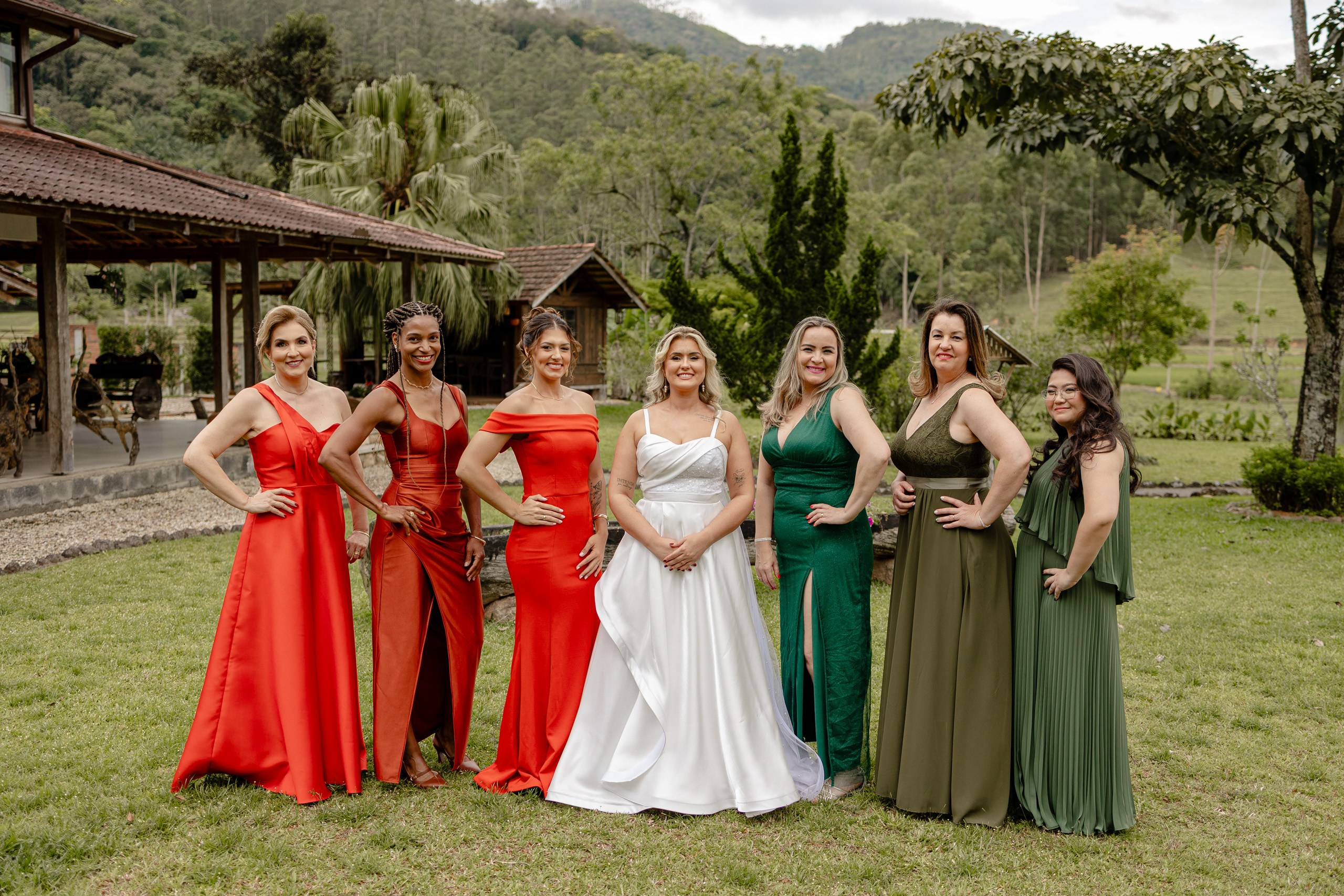 Fotógrafo de casamento em Santa Catarina; noiva de vestido branco posando ao lado de madrinhas com vestidos em tons de vermelho e verde em área externa ao ar livre.