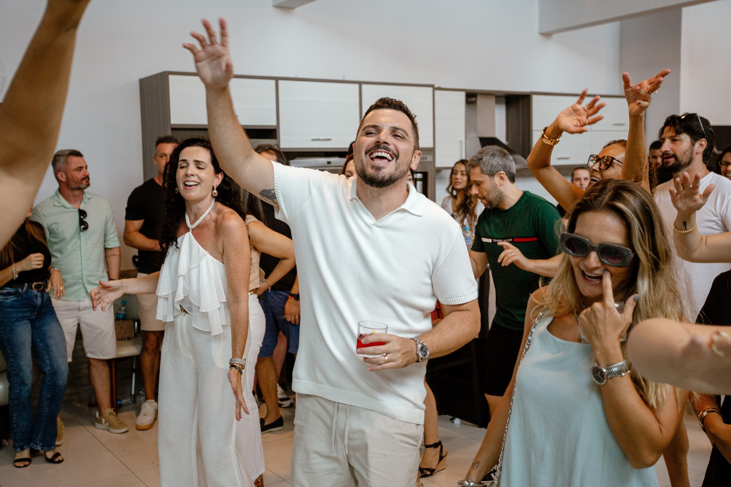 Fotografia de cobertura de festa de aniversário em Itajaí capturando o marido da aniversariante a sorrir e a celebrar alegremente com os braços no ar rodeado por convidados.