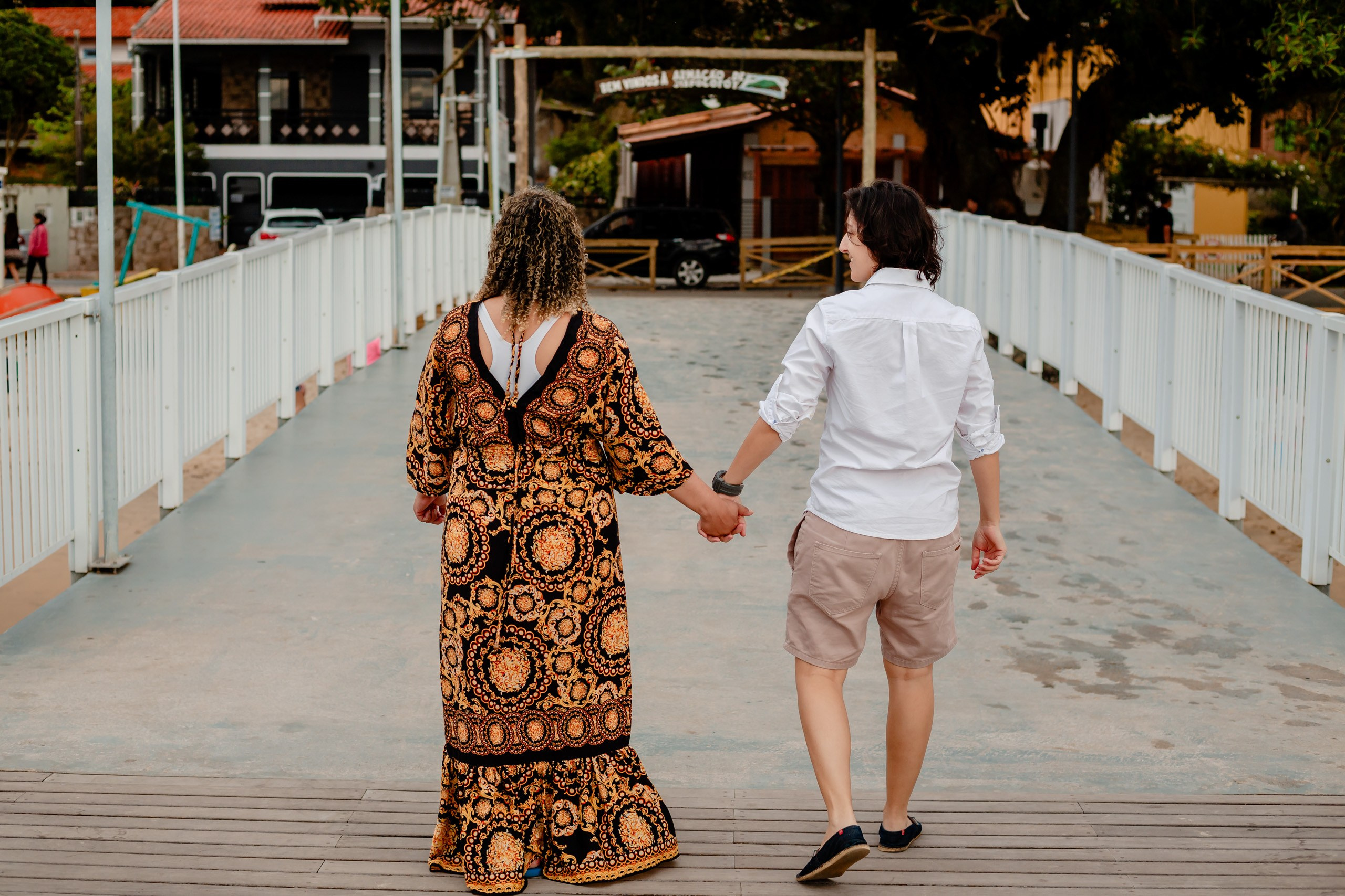 Fotógrafo ensaio gestante em Penha; plano de costas do casal caminhando de mãos dadas em uma passarela de madeira, em um registro espontâneo e afetuoso.