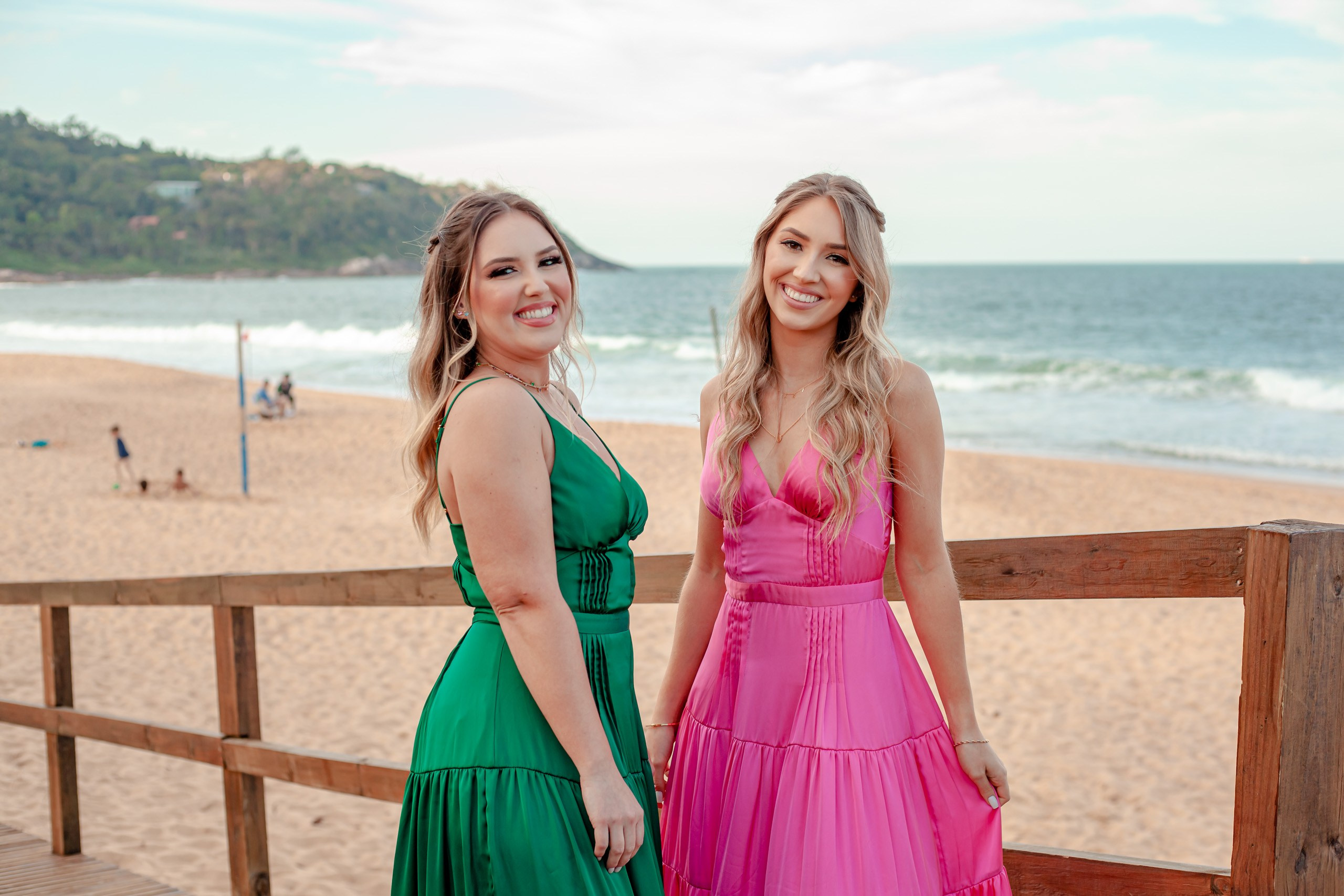 Fotógrafo festa de formatura em Balneário Camboriú; retrato de meio corpo das duas formandas sorridentes na passarela com vista para a praia e o mar no Bali Bistro Beach.