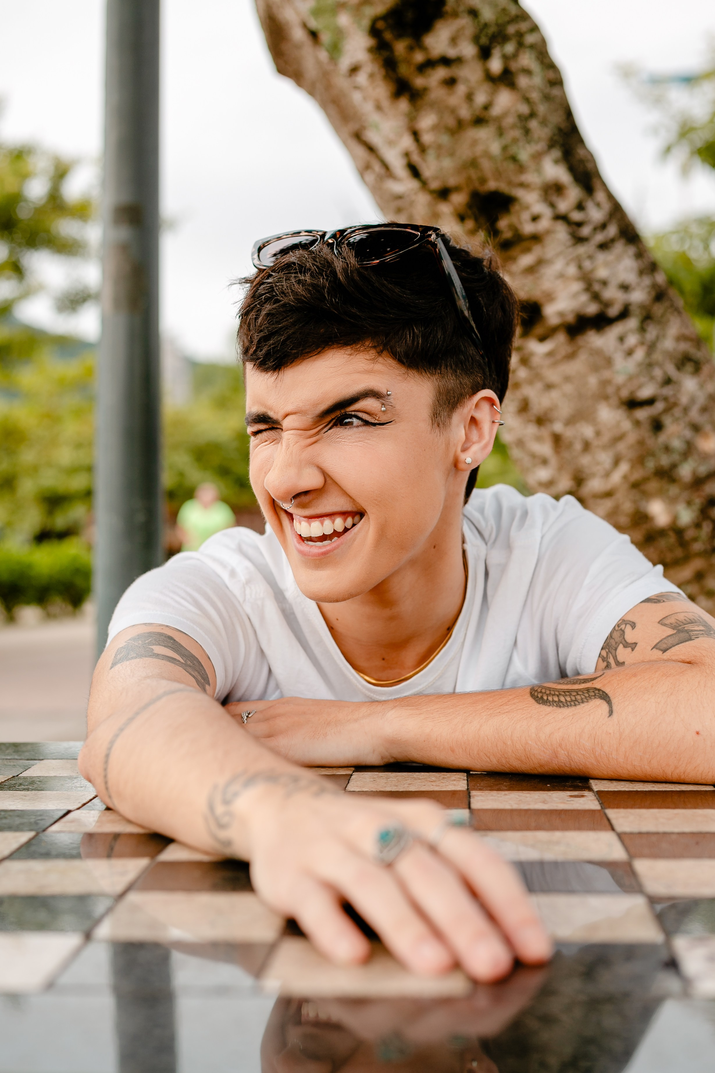 Fotógrafo de ensaio em Balneário Camboriú; retrato dinâmico e divertido de jovem com cabelo curto escuro, piercings, tatuagens e t-shirt branca, sorridente e apoiada numa mesa de xadrez ao ar livre.