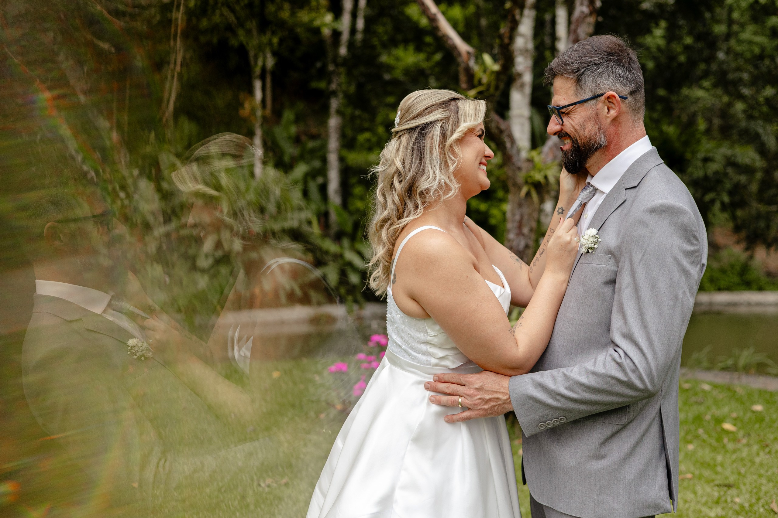 Fotógrafo de casamento em Balneário Camboriú; retrato romântico e descontraído da noiva a ajeitar a gravata do noivo, com o casal sorrindo um para o outro em um jardim arborizado ao ar livre.