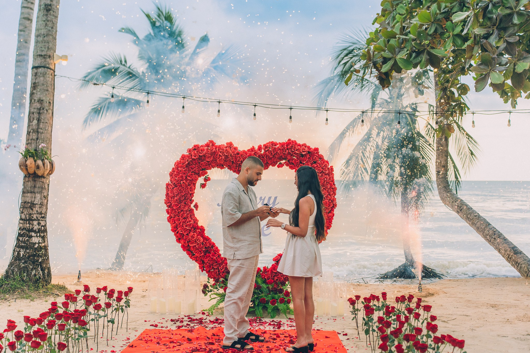 Man proposing on one knee to his partner on a decorated Pattaya beach with red roses and rose petals.