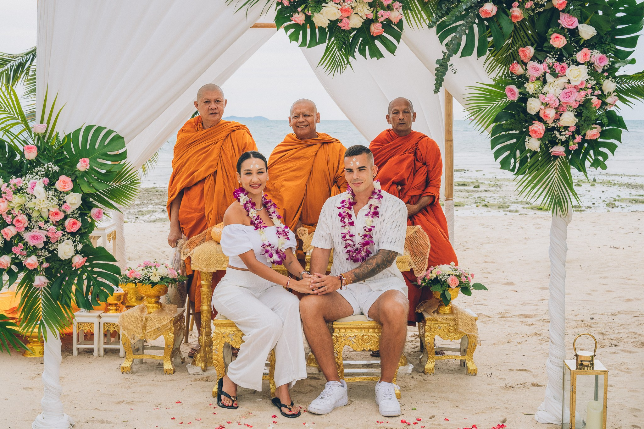 Traditional Buddhist beach wedding ceremony in Koh Samui, Thailand, with a bride and groom seated in front of three Buddhist monks, surrounded by