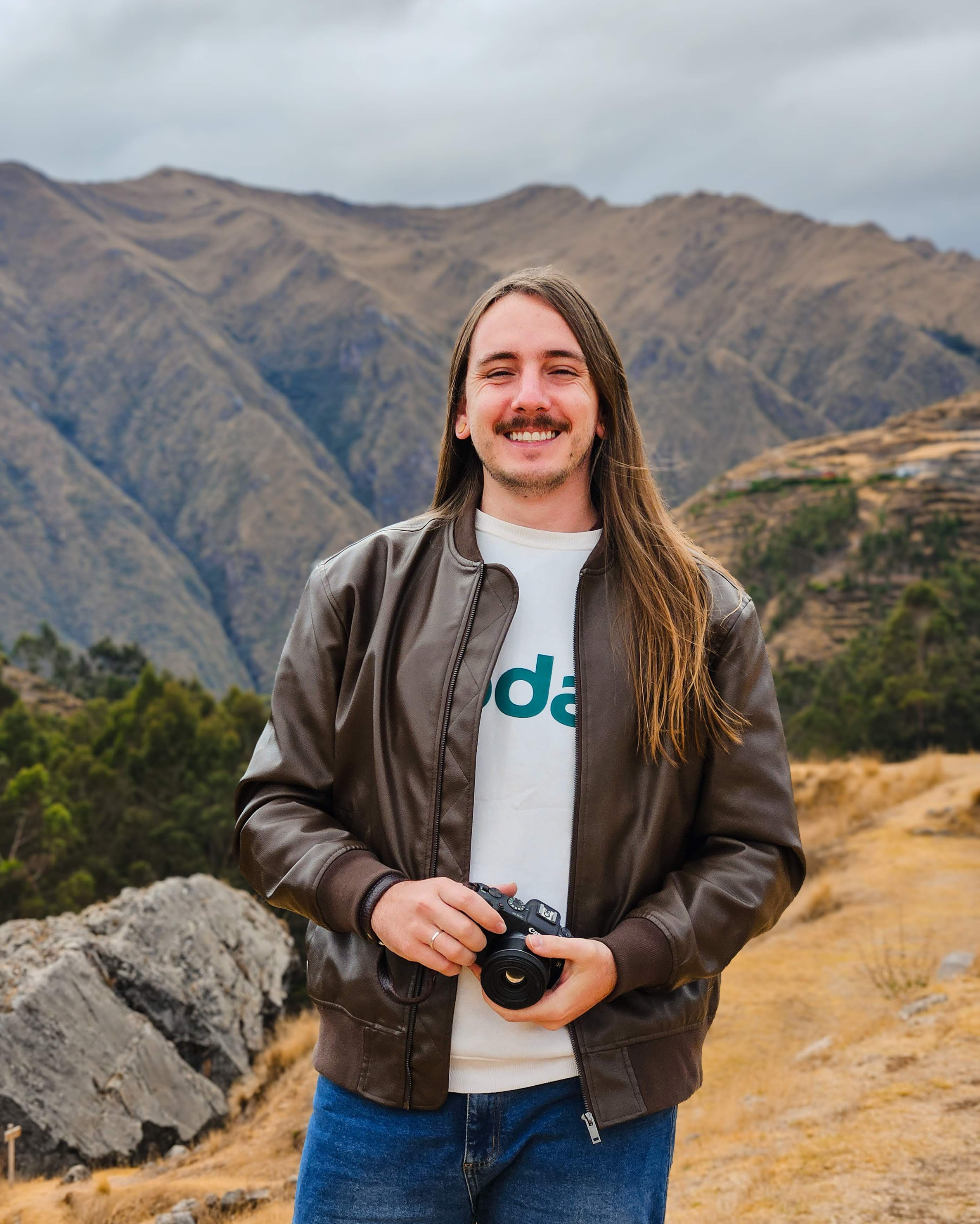 Fotógrafo segurando câmera com jaqueta de couro em paisagem montanhosa durante viagem.