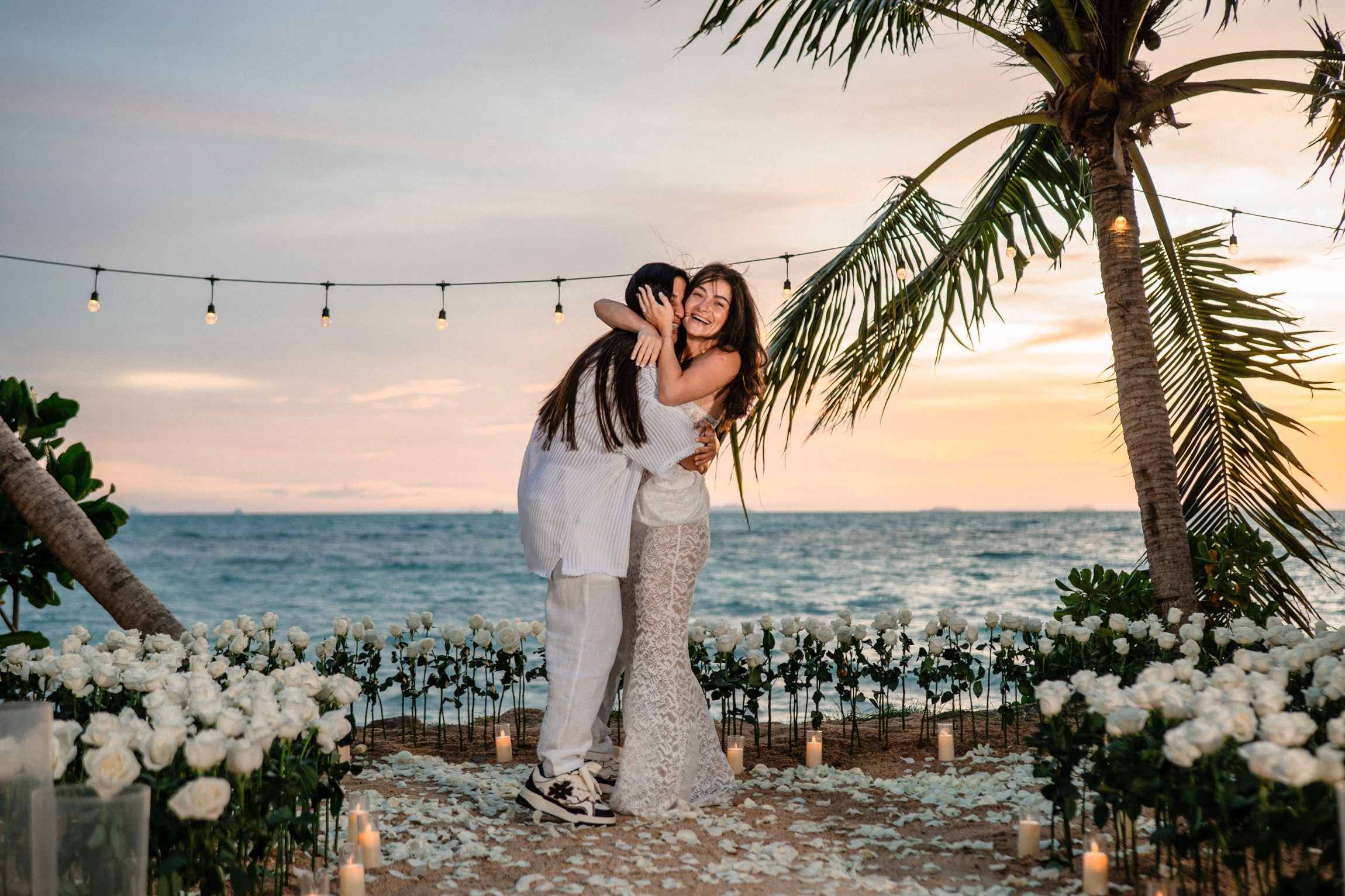 Happy couple of 2 lady celebrating their engagement on a romantic Koh Samui beach at sunset with white roses, candles and ocean views.