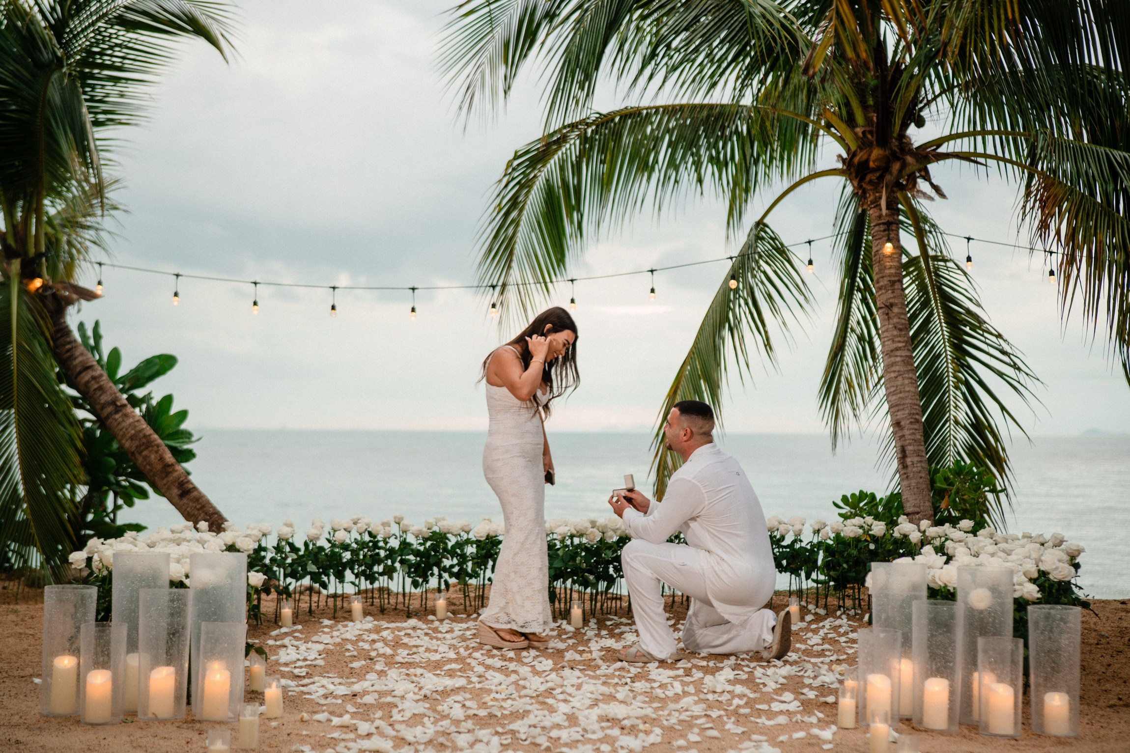 Beach marriage proposal in Koh Samui with a man on one knee, white roses, candles and a romantic ocean view.