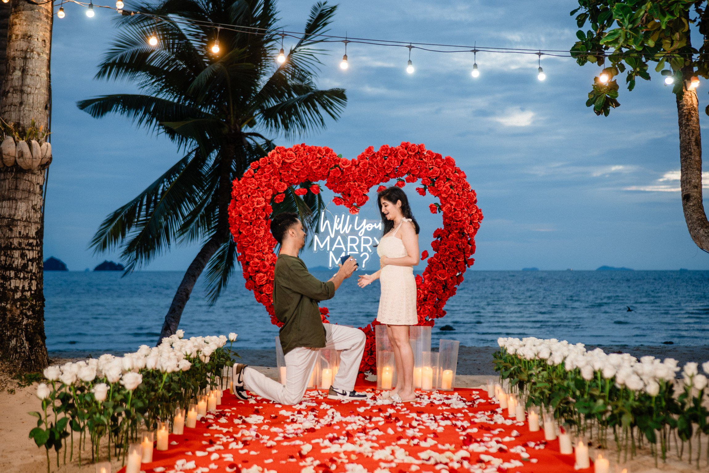 Romantic beach marriage proposal in Koh Samui with a red heart-shaped rose arch, candles and an ocean backdrop.