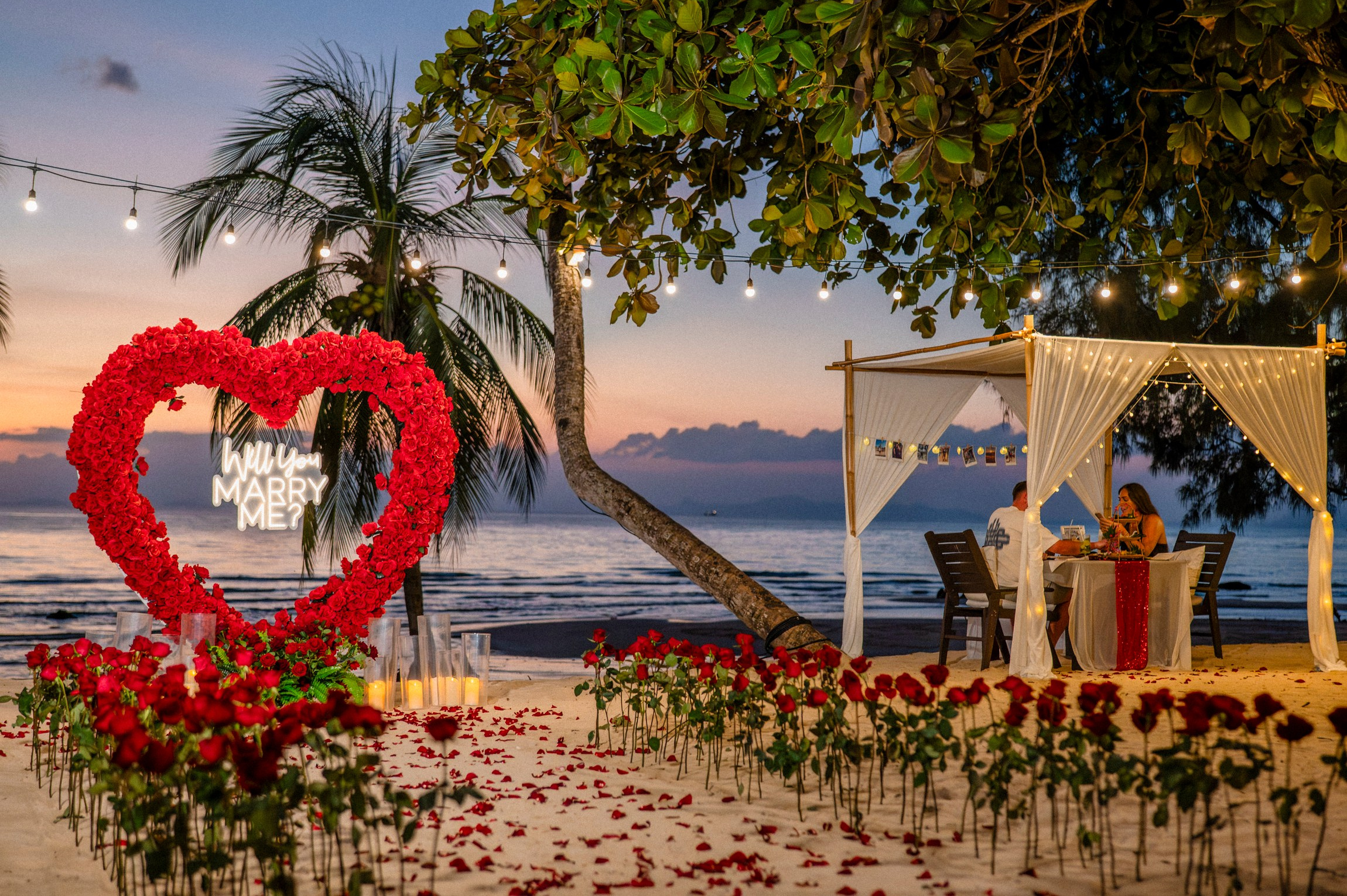 Romantic beach proposal setup in Pattaya with a red floral heart, rose petals, candles and a decorated seaside table.