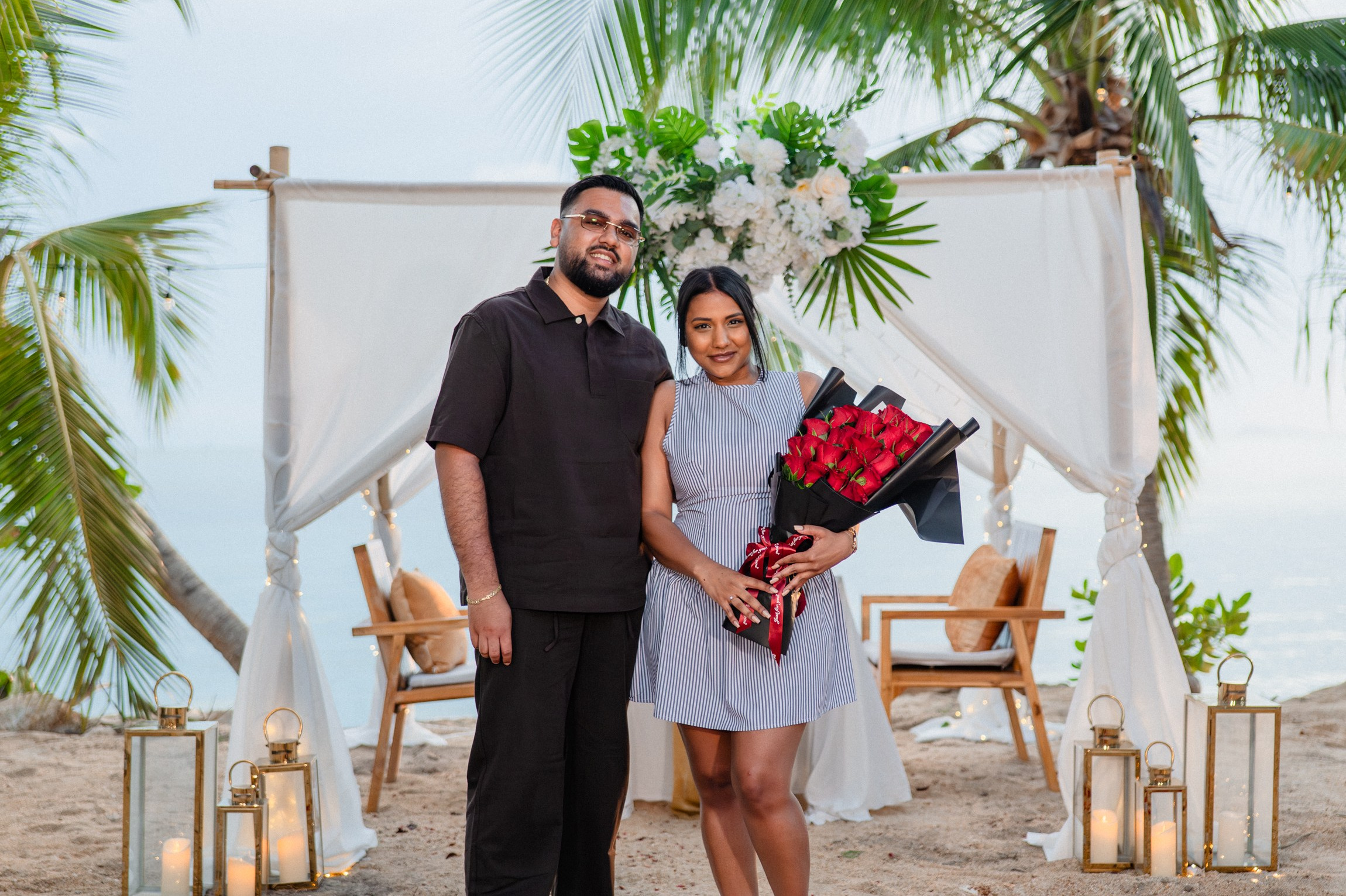Newly engaged couple posing with a bouquet of red roses beside a romantic beach proposal setup in Pattaya.