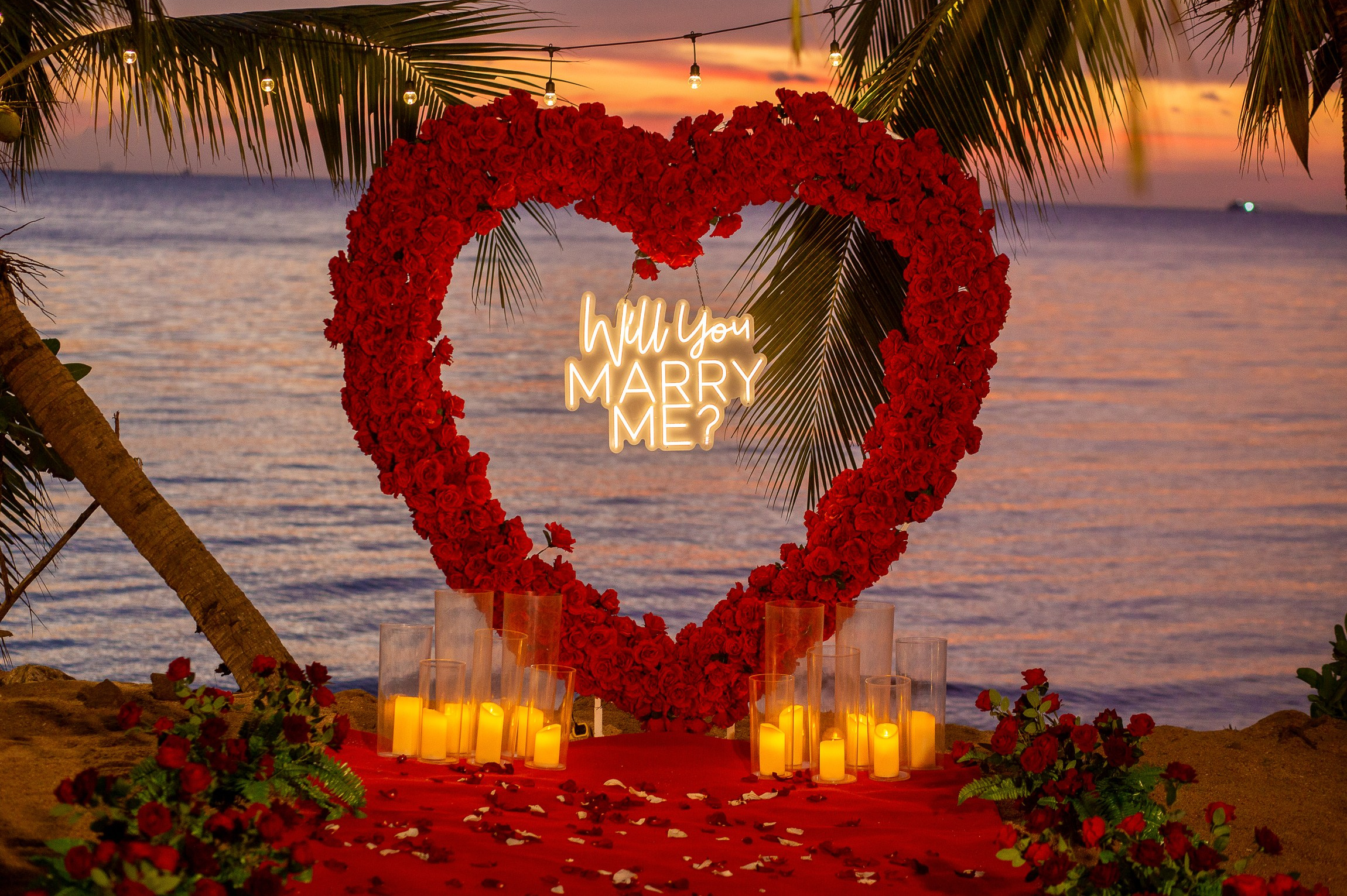 Heart-shaped arch covered with red roses and a glowing “Will You Marry Me?” sign on the beach in Pattaya at sunset.