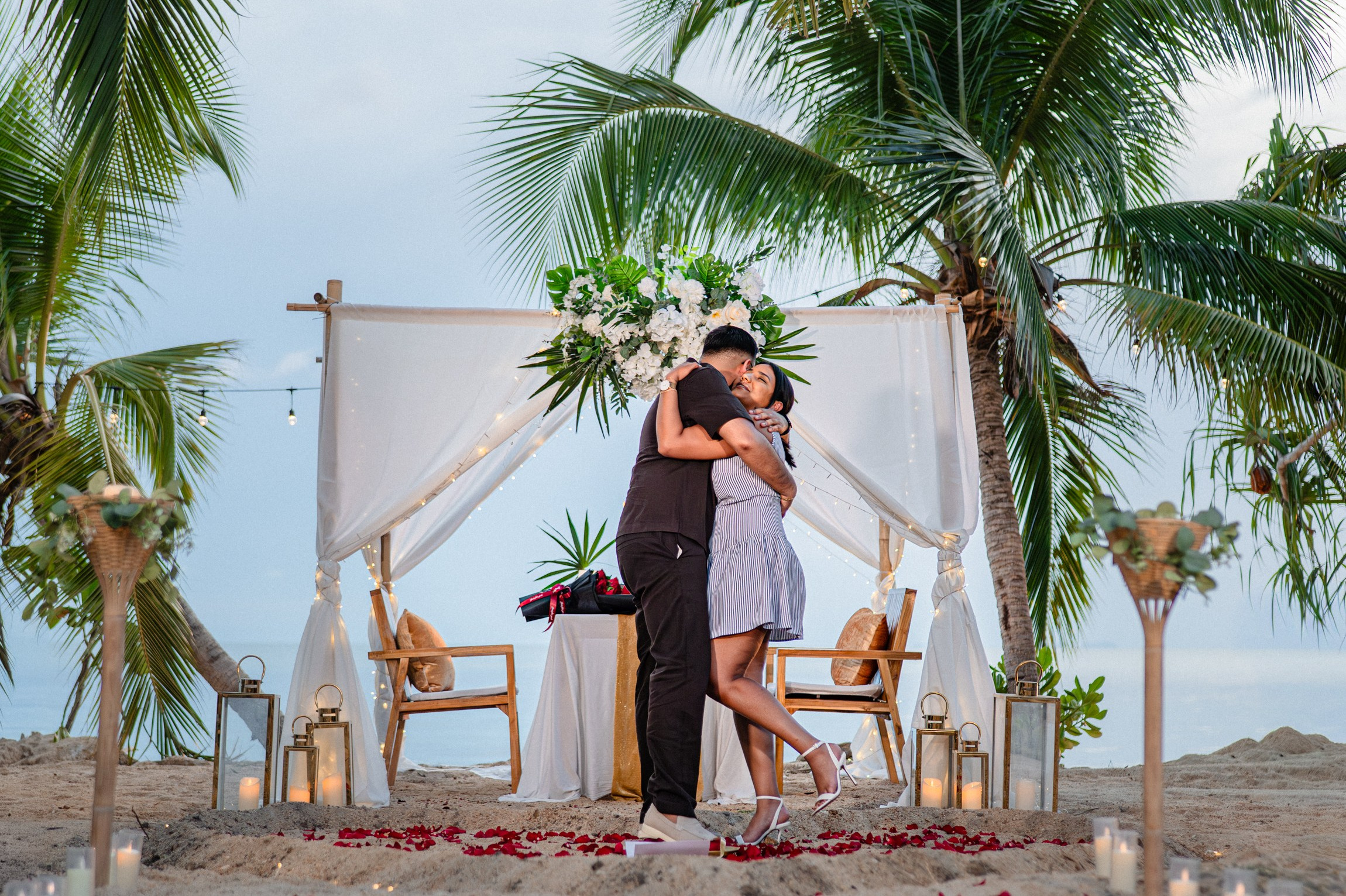 Happy engaged couple embracing beside an elegant tropical beach proposal setup in Pattaya.