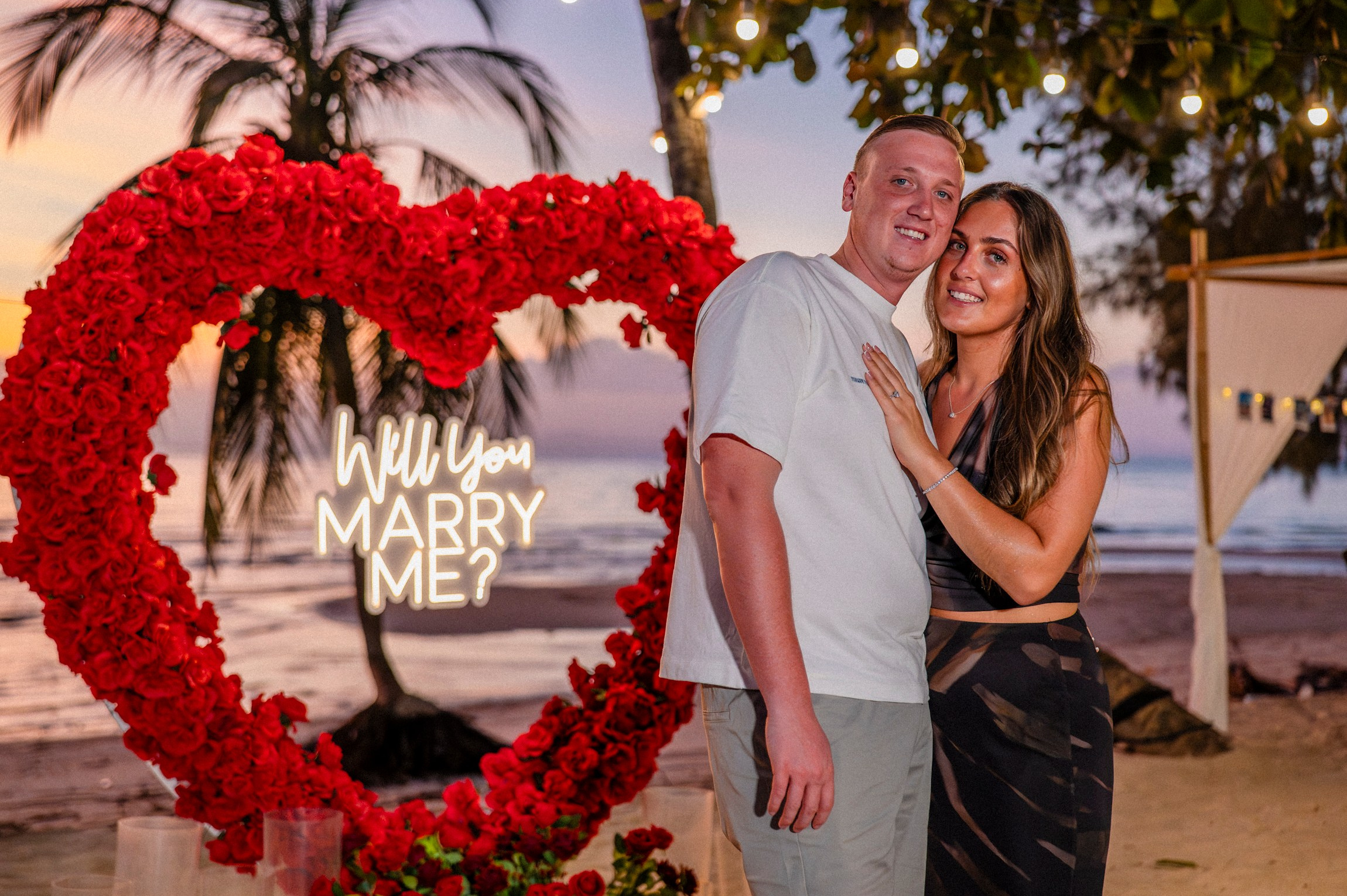 Happy couple posing beside a red rose heart decoration after a romantic marriage proposal in Pattaya.
