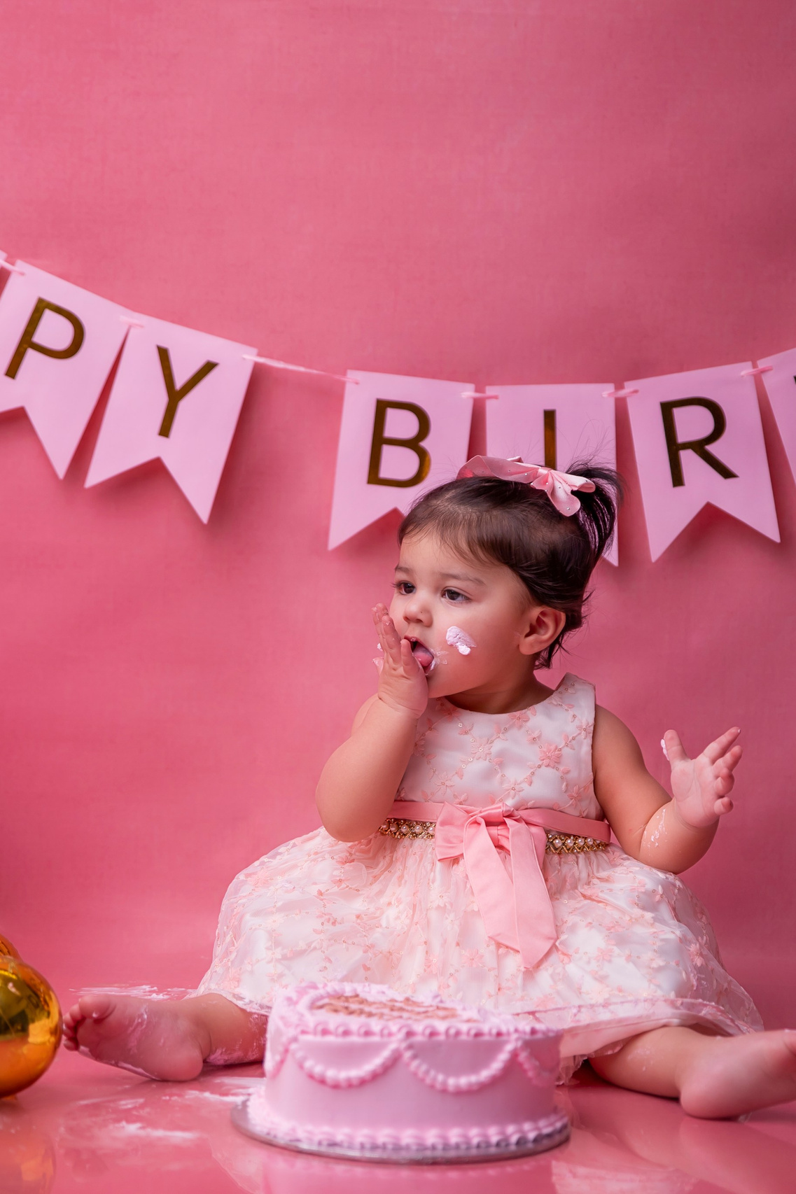 An adorable little girl/baby with a pink frock/dress and pink background with a  happy birthday banner/streamer behind her.cake smash. birthday bash. birthday photo.  baby photography. peshawar. faryal's baby photography. baby pictures. newborn. newborn picture. newborn pictures. newborn photography.
