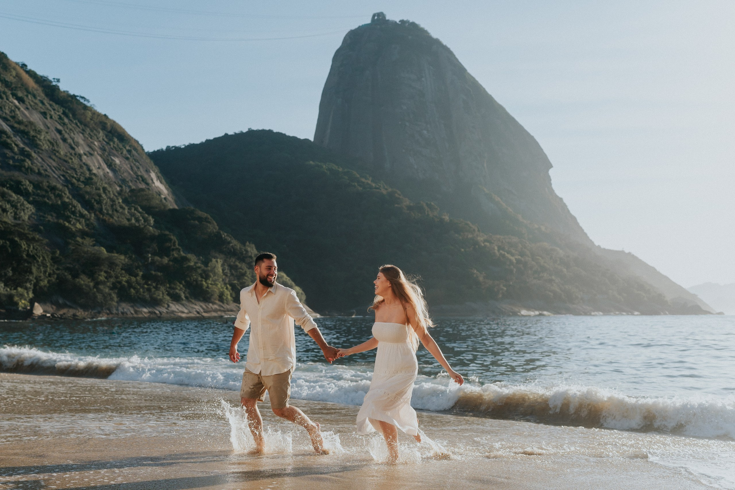 Casal correndo e sorrindo em ensaio pré-wedding ao ar livre com vista para o Pão de Açúcar, romântico destination wedding no Rio de Janeiro.