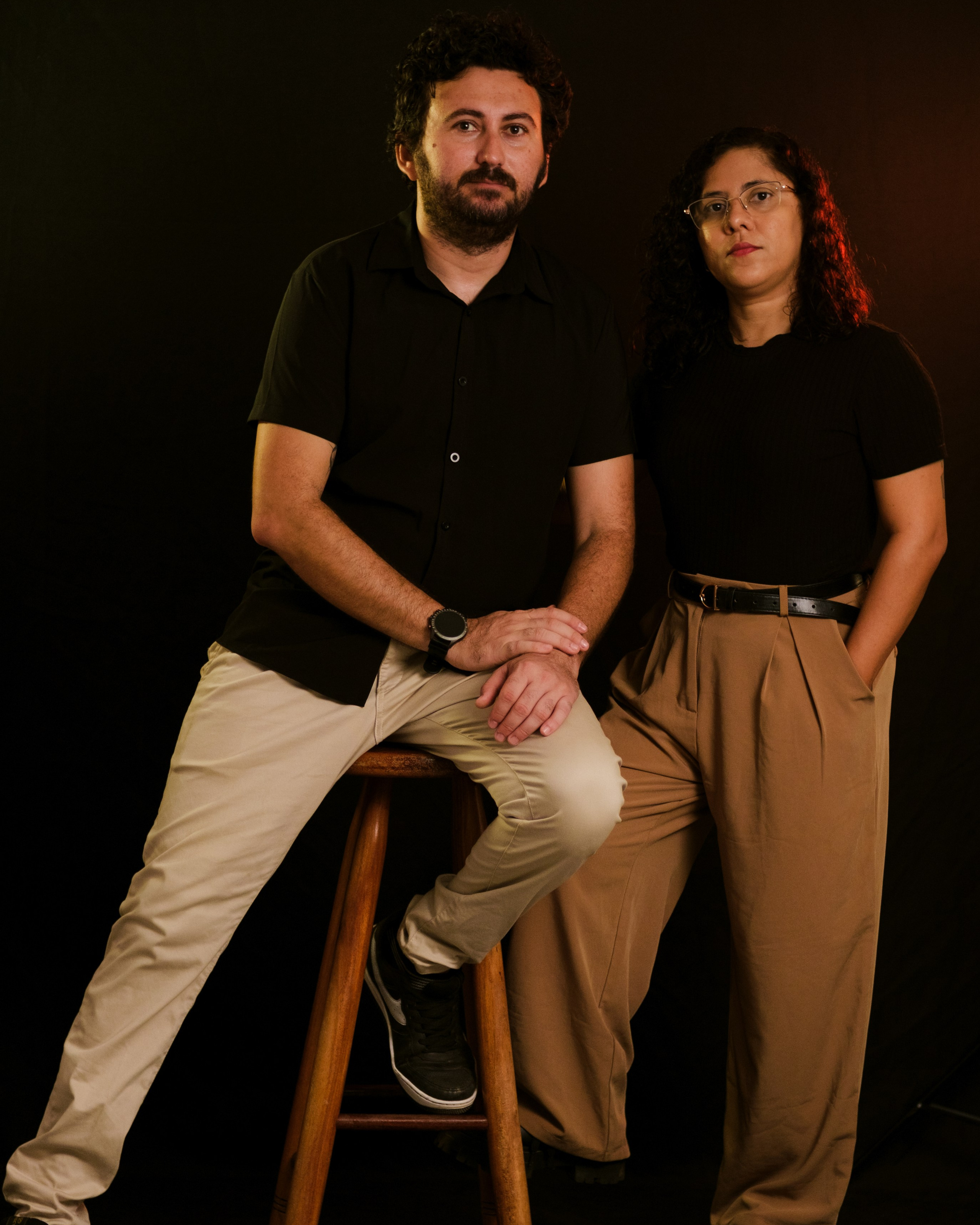 casal de fotógrafos em fotos de estúdio ele está sentado em um banco de madeira e ela está em pé perto dele com as mãos nos bolsos, os dois estão olhando para a câmera