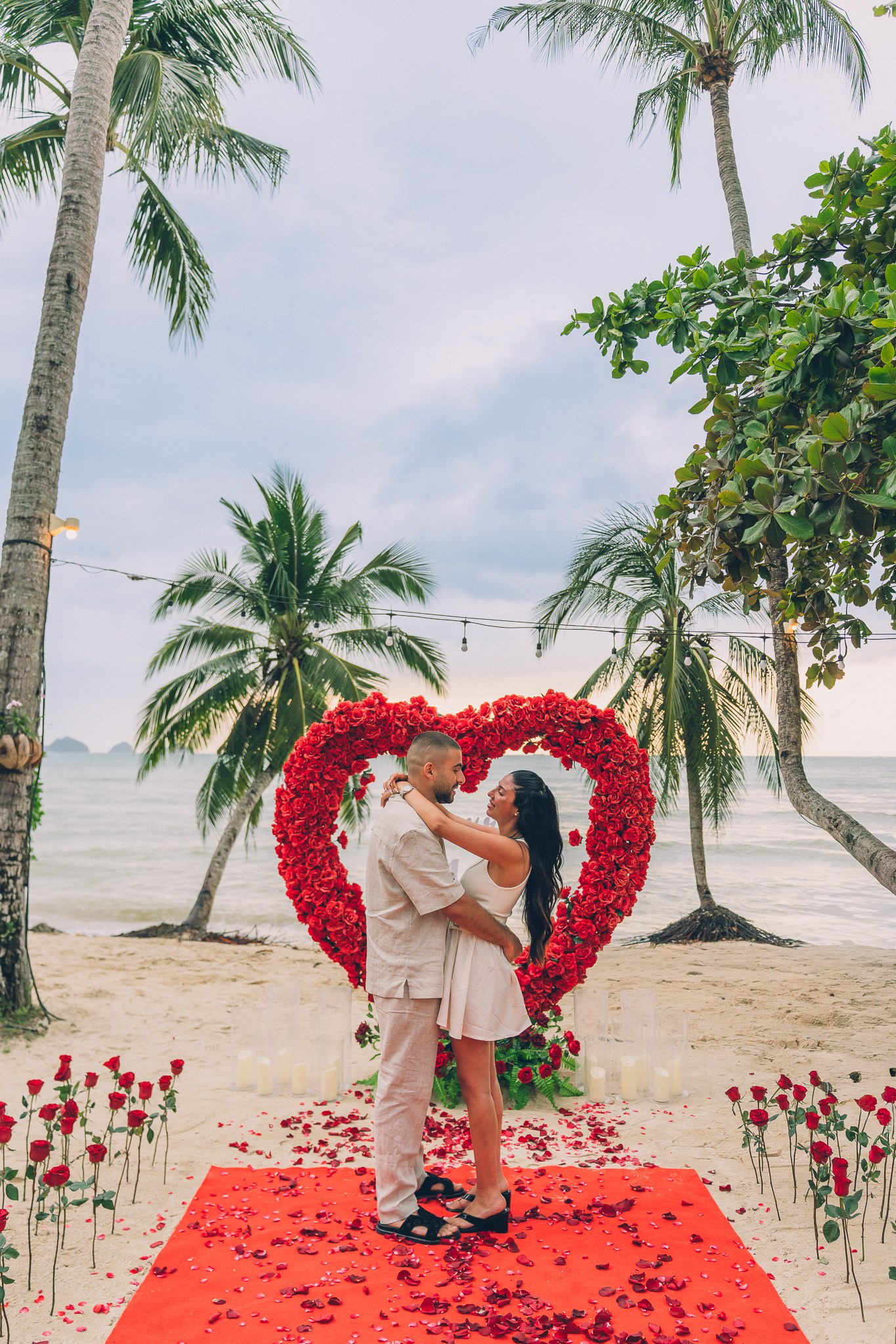 Newly engaged couple embracing in front of a red rose heart arch on a romantic Pattaya beach.