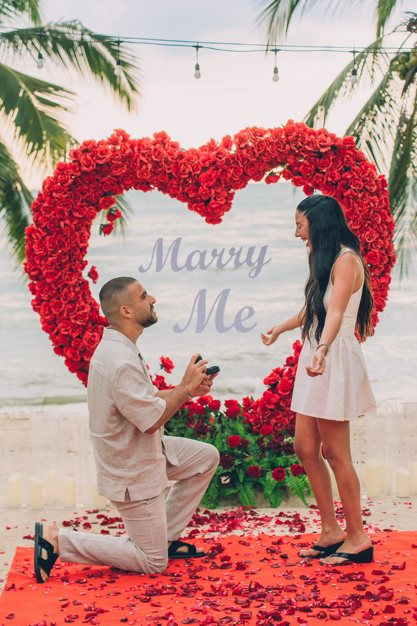 Man proposing on one knee in front of a large red rose heart with “Marry Me” decoration on a Pattaya beach.
