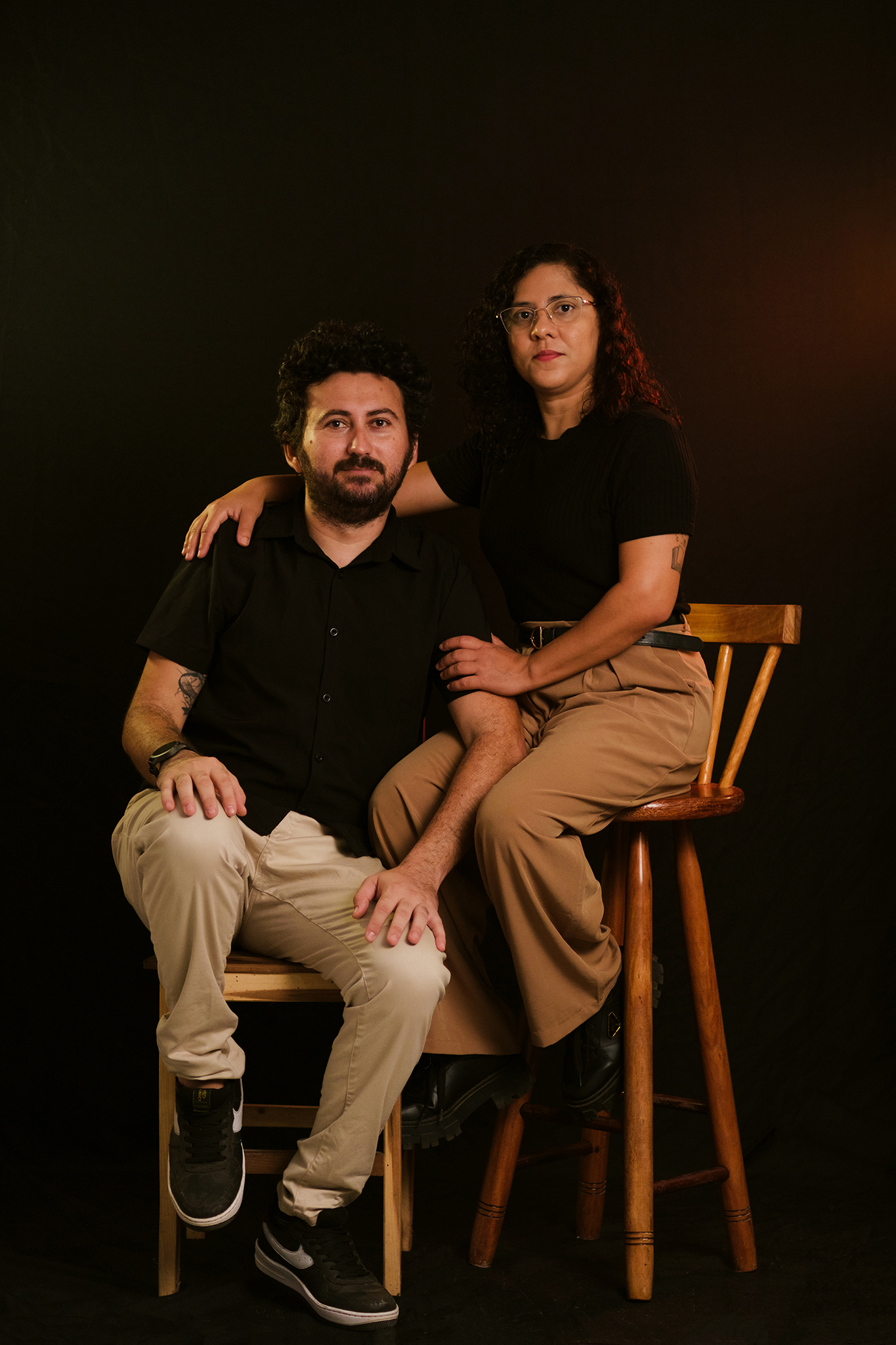 casal de fotógrafos sentados em cadeiras de madeira, ela encosta a mão no ombro dele e os dois estão olhando para a câmera