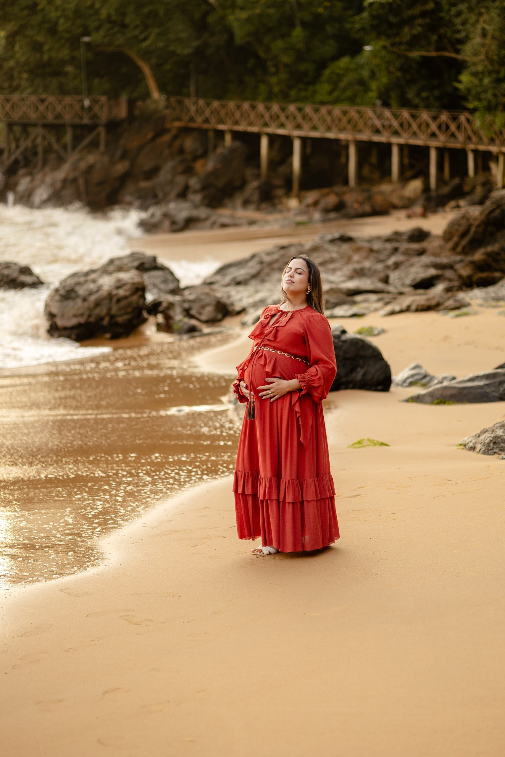 Fotógrafo de eventos em Itajaí; retrato solo e delicado de gestante com vestido longo vermelho, acariciando a barriga na areia da praia, tendo rochas e uma passarela de madeira ao fundo.