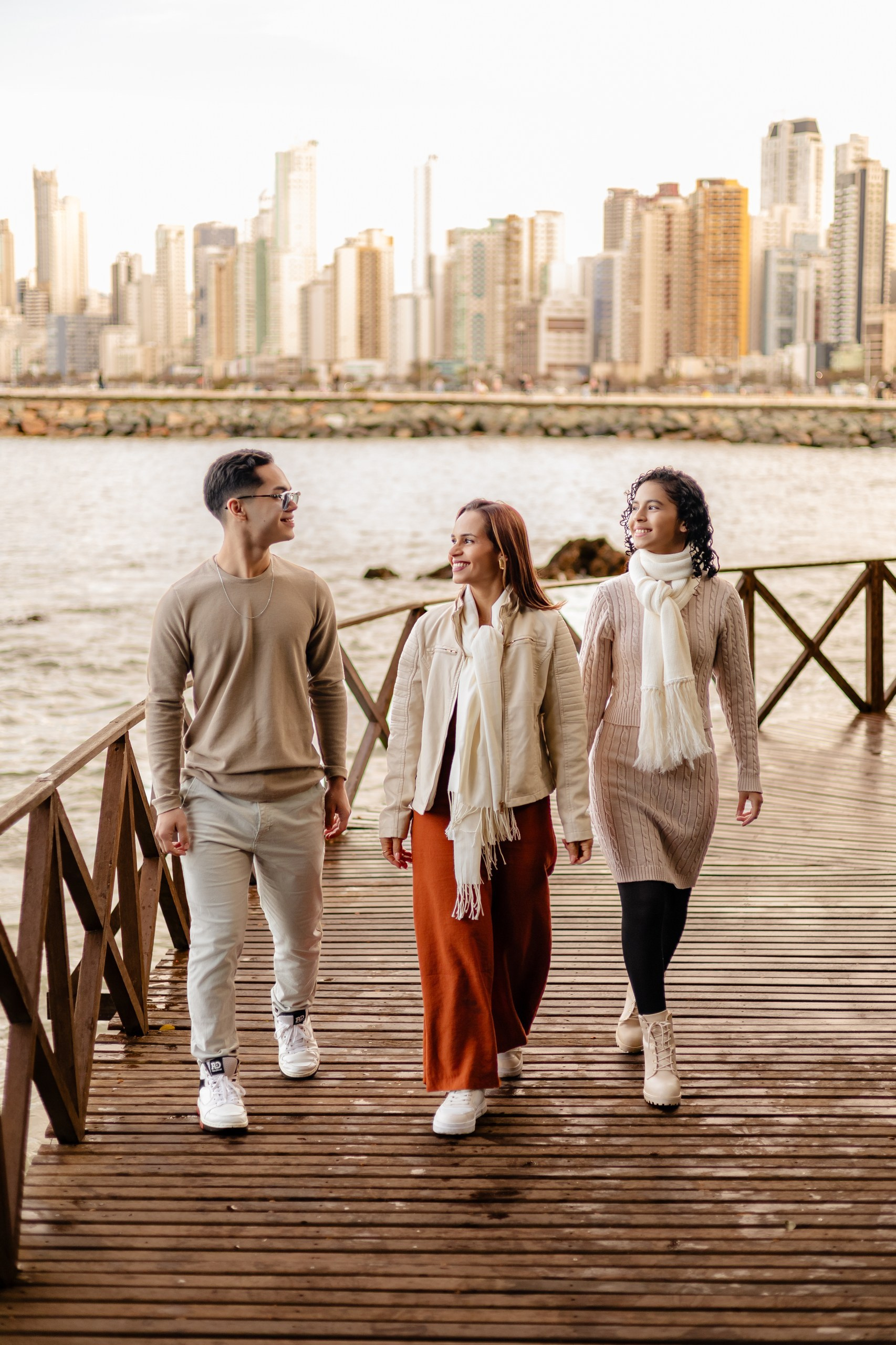Fotógrafo de eventos em Itajaí; retrato dinâmico e natural de três pessoas caminhando juntas numa passarela de madeira, com o icônico skyline da cidade e o mar ao fundo.